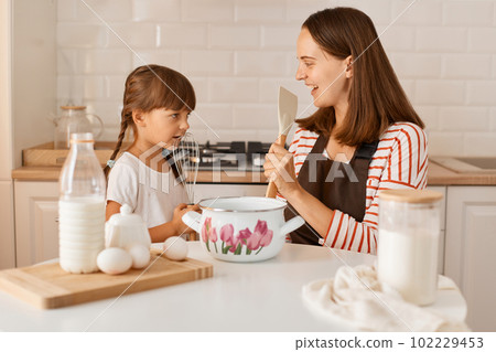 Extremely happy excited young adult woman cooking with her daughter at home, having fun together, singing with kitchen equipment, enjoying spending time together. Extremely happy excited young adult woman cooking with her daughter at home, having fun together, singing with kitchen equipment, enjoying spending time together. 102229453