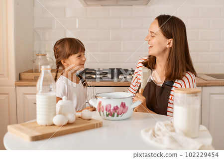 Horizontal shot of delighted young adult woman cooking with her daughter in the kitchen, having fun together, laughing, looking at each other with happy facial expression. Horizontal shot of delighted young adult woman cooking with her daughter in the kitchen, having fun together, laughing, looking at each other with happy facial expression. 102229454