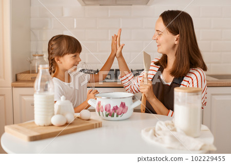 Image of happy mother baking together with her daughter, sitting at table in the kitchen, kid giving five to mommy, smiling, enjoying time while making homemade pastry. Image of happy mother baking together with her daughter, sitting at table in the kitchen, kid giving five to mommy, smiling, enjoying time while making homemade pastry. 102229455