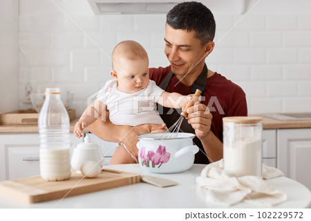 Horizontal shot of positive good looking joyful dad sitting at table with toddler daughter and cooking together in kitchen, mixing dough for bakery with tiny baby's hand. 102229572