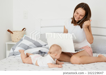 Indoor shot of happy joyful woman sitting on bed with her infant daughter and working online, holding laptop and talking cell phone, clenched fist, celebrating success. 102229647