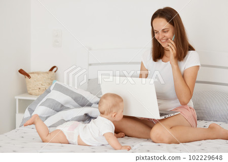 Portrait of young adult mother posing with baby while talking on phone and working on laptop at home, woman wearing white casual style t shirt while sitting on bed with baby. 102229648