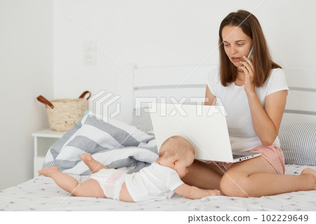 Horizontal shot of winsome beautiful busy woman sitting on bed with her infant daughter and working online, holding laptop, talking on mobile phone with partner, playing with baby. 102229649