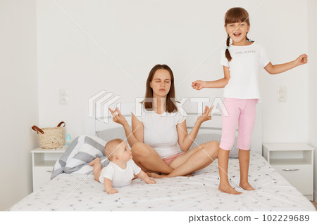 Mother doing morning exercises in yoga pose while sitting on bed in light room, her little daughters playing near at home, happy little girl standing with happy expression near mommy. 102229689