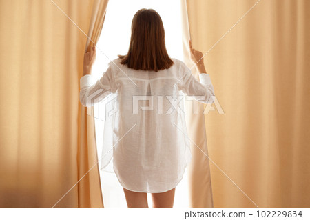 Back side portrait of dark haired female wearing cotton white shirt open curtains, standing near window, woman with beautiful perfect body posing backwards at home. 102229834