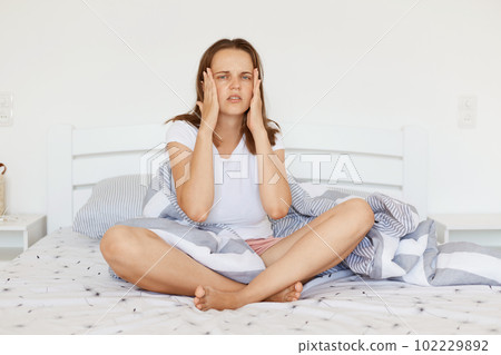 Portrait of young adult woman wearing white casual t shirt, sitting with crossed legs on bed in bedroom, having terrible headache, keeping hands on her temples. 102229892