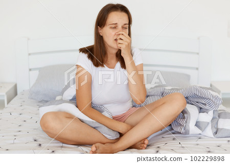 Young adult dark haired woman wearing white casual t shirt sneezing, catching a cold, suffering from fever, keeping hand on nose, posing in light bedroom. Young adult dark haired woman wearing white casual t shirt sneezing, catching a cold, suffering from fever, keeping hand on nose, posing in light bedroom. 102229898