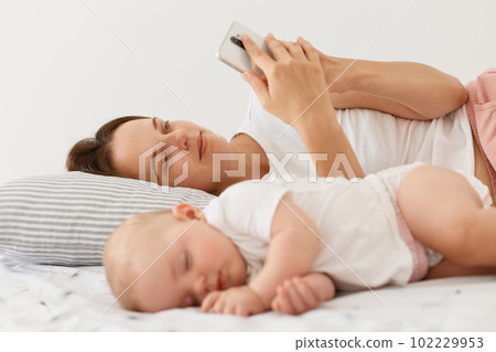 Portrait of sleeping toddler daughter wearing white attire lying near mother, woman using smart phone when wakes up and checking emails or social networks. 102229953