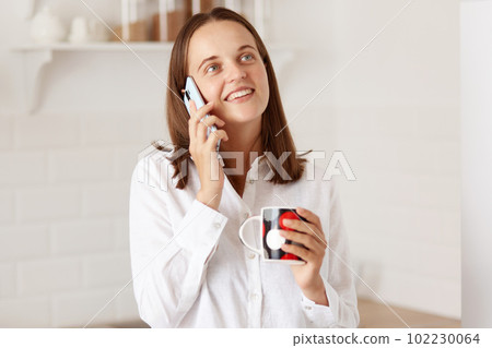 Indoor shot of beautiful girl talking on the mobile phone, holding a cup of coffee or tea, looking away and smiling while posing with the kitchen set on background. 102230064