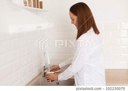 Side view portrait of dark haired woman washing dish or pouring cup with fresh drink water at kitchen faucet, female wearing white casual style shirt, posing in kitchen. 102230070
