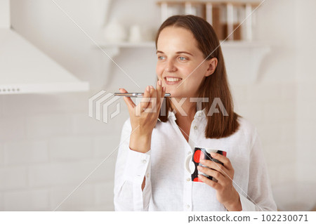 Indoor shot of attractive young adult woman making a voice message with smart phone while relaxing at home and drinking hot tea or coffee, standing in light kitchen. Indoor shot of attractive young adult woman making a voice message with smart phone while relaxing at home and drinking hot tea or coffee, standing in light kitchen. 102230071