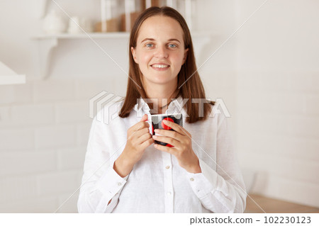 Indoor shot of smiling pretty woman having a cup of coffee in kitchen, standing with pleasant smile, enjoying hot tea in the morning after breakfast, looking at camera with positive. Indoor shot of smiling pretty woman having a cup of coffee in kitchen, standing with pleasant smile, enjoying hot tea in the morning after breakfast, looking at camera with positive. 102230123