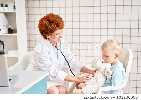Female Pediatrician listening to breathing in the lungs and heartbeat with stethoscope. Portrait of adorable little boy visiting doctor. Female Pediatrician listening to breathing in the lungs and heartbeat with stethoscope. Portrait of adorable little boy visiting doctor. 102230360