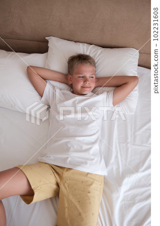 Handsome teenager lies in bed in a white t-shirt in the bedroom at home and looks out the window. Handsome teenager lies in bed in a white t-shirt in the bedroom at home and looks out the window. 102231608