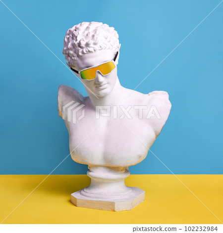 Antique male statue bust wearing stylish, trendy sunglasses against blue yellow background. Summer vibes Antique male statue bust wearing stylish, trendy sunglasses against blue yellow background. Summer vibes 102232984