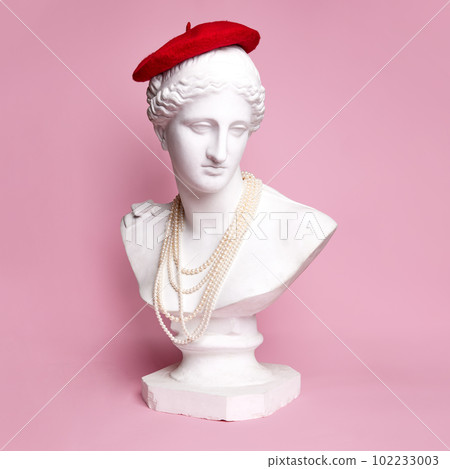 Antique statue bust of pretty woman wearing red beret and pearl necklace against pink background. Parisian look 102233003