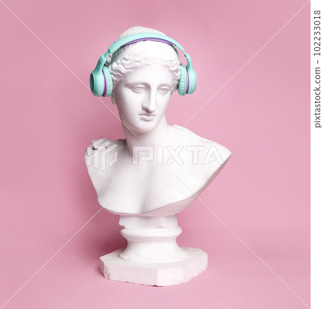 Antique statue bust wearing blue headphones against pink background. Music lifestyle, technologies, relaxation Antique statue bust wearing blue headphones against pink background. Music lifestyle, technologies, relaxation 102233018