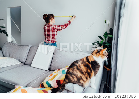 Young woman doing measuring with a measure tape on the wall. Girl wants to put a picture on the wall at home. Housekeeping work. Doing repair herself. DIY, gender equality in work concept. Young woman doing measuring with a measure tape on the wall. Girl wants to put a picture on the wall at home. Housekeeping work. Doing repair herself. DIY, gender equality in work concept. 102237399