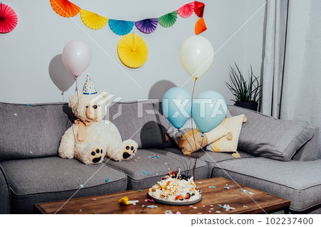 Messy table in living room after party. Broken birthday cake, confetti, balloons and decoration. Big teddy bear sitting on the sofa. Selective focus 102237400