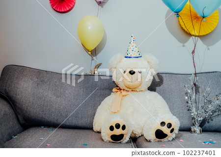 Big Teddy bear with balloons in party cap sitting on the gray sofa in decorated room. Birthday or festive holiday background. Selective focus, copy space 102237401