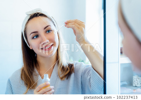 Young woman in bathrobe looking in the mirror, holding dropper with hyaluronic acid serum close to face and smiling. Skin hydrating. Cosmetic spa procedures. Beauty self-care at home. Selective focus. Young woman in bathrobe looking in the mirror, holding dropper with hyaluronic acid serum close to face and smiling. Skin hydrating. Cosmetic spa procedures. Beauty self-care at home. Selective focus. 102237413