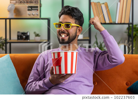 Indian man sits on couch eating popcorn and watching interesting TV serial sport game online at home Indian man sits on couch eating popcorn and watching interesting TV serial sport game online at home 102237603