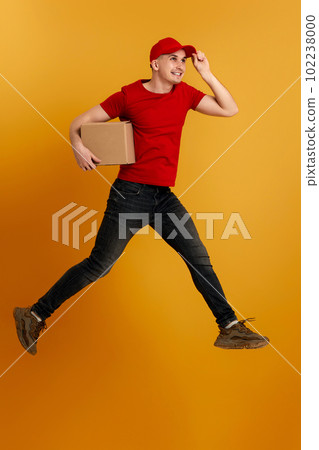 Delivery man in red cap, t-shirt holding cardboard box and jumping Delivery man in red cap, t-shirt holding cardboard box and jumping 102238000
