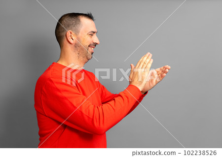 Bearded hispanic man in profile wearing a red jumper feeling happy, smiling and clapping with grateful emphasis for the great show he just witnessed. Isolated on gray studio background Bearded hispanic man in profile wearing a red jumper feeling happy, smiling and clapping with grateful emphasis for the great show he just witnessed. Isolated on gray studio background 102238526