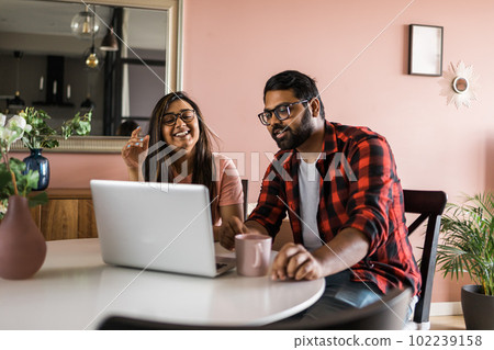 Happy indian family couple cuddle at desk make video call to friends using laptop webcam. Loving young spouses look at computer screen waving hands in good mood greeting parents communicating online 102239158