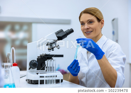 Scientist working in scientific laboratory, holding test tube with blue liquid, using microscope 102239478