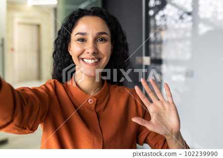 Beautiful business woman with curly hair wearing looking at smartphone camera, waving her hand in greeting gesture, talking with friends and colleagues, using video call app. Beautiful business woman with curly hair wearing looking at smartphone camera, waving her hand in greeting gesture, talking with friends and colleagues, using video call app. 102239990