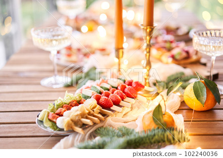 Christmas dinner feast. A small table is served with snacks, bruschettas, and canapes. A decorated dining table with champagne glasses, candles and christmas tree an garland in background 102240036