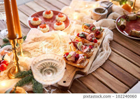 Christmas dinner feast. A small table is served with snacks, bruschettas, and canapes. A decorated dining table with champagne glasses, candles and christmas tree an garland in background 102240060
