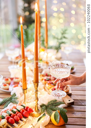 Hand with a glass of champagne in the foreground. A small table is served with snacks, bruschettas, and canapes. A decorated dining table with champagne glasses, candles and christmas tree an garland 102240068