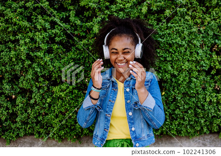 Multiracial girl enjoying music in headphones, standing in front of green bush, portrait. 102241306