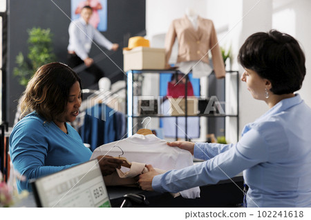 African american woman giving multiple shirts to worker, discussing fabric before buying it in clothing store. Stylish customer shopping for casual wear, purchasing new fashion collection 102241618