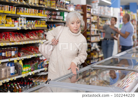 Mature woman choosing frozen food in supermarket. Young woman purchasing goods in grocery store Mature woman choosing frozen food in supermarket. Young woman purchasing goods in grocery store 102244220