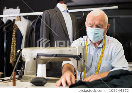 Portrait of elderly tailor in protective mask working on sewing machine 102244372