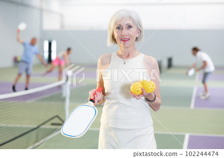 Smiling sporty elderly woman standing in indoor pickleball court Smiling sporty elderly woman standing in indoor pickleball court 102244474