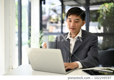 Smart Asian businessman sipping coffee while using laptop computer at his desk. 102249597