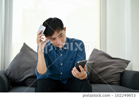 Confused Asian man sits on a sofa, having a problem with his mobile banking application. Confused Asian man sits on a sofa, having a problem with his mobile banking application. 102249653
