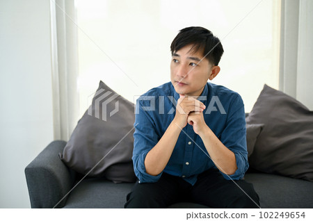 Thoughtful Asian man sits on a sofa, pondering and looking out the window. Thoughtful Asian man sits on a sofa, pondering and looking out the window. 102249654
