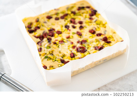 Removing cranberry pistachio fudge from a square cheesecake pan lined with parchment. 102250213