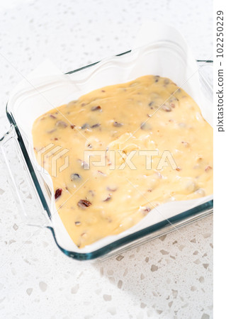 Pouring fudge mixture into the baking pan lined with parchment paper to prepare white chocolate cranberry pecan fudge. Pouring fudge mixture into the baking pan lined with parchment paper to prepare white chocolate cranberry pecan fudge. 102250229