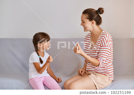 Side view of woman professional physiotherapist working on speech defects with cute child girl while sitting on gray sofa, kid with braids having language lesson for improving speaking. 102251199