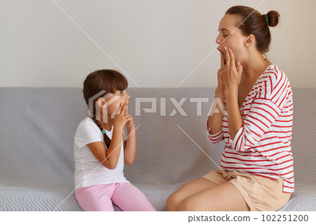 Side view of young adult Caucasian language therapist teaching little kid sounds pronunciation, working on speech defects or difficulties with small child girl indoors while sitting on sofa. 102251200
