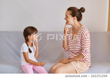 Portrait of woman and little girl having lesson for improving sounds or language difficulties, female child sitting indoor on comfortable sofa and practicing articulation. 102251204