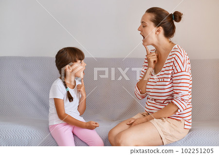 Profile shot of woman physiotherapist having lesson with small child girl for correct articulation, people sitting on sofa, small kid improving speaking. 102251205