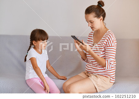 Professional female physiotherapist working with small child girl, sitting on sofa with mobile phone in hands, finds new online lesson in her gadget for training. 102251210