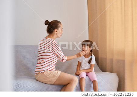 Indoor shot of young adult female speech therapist working with little girl, people wearing casual style clothing, sitting on sofa near window and demonstrating how to pronounce sounds and words. Indoor shot of young adult female speech therapist working with little girl, people wearing casual style clothing, sitting on sofa near window and demonstrating how to pronounce sounds and words. 102251225
